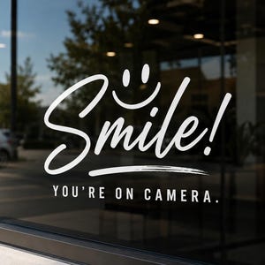 May include: A white vinyl decal applied to a glass surface featuring the word Smile in a large cursive script with a smiley face icon above it and the text YOU'RE ON CAMERA in a smaller sans-serif font positioned directly underneath.