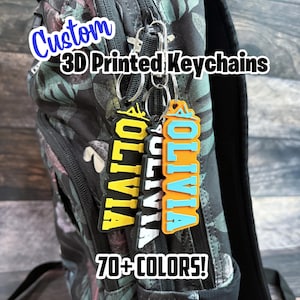 May include: Custom 3D printed keychains with the name "Olivia" in yellow and orange with blue accents. The keychains are attached to a backpack. The image also includes the text "70+ COLOURS!"