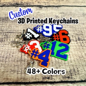 Puede incluir: Una colección de llaveros personalizados impresos en 3D con varios números, incluidos 1, 3, 4, 6, 9 y 12, en varios colores como azul, naranja, verde, blanco y negro, cada uno unido a un anillo de llavero con cierre de mosquetón de metal plateado.