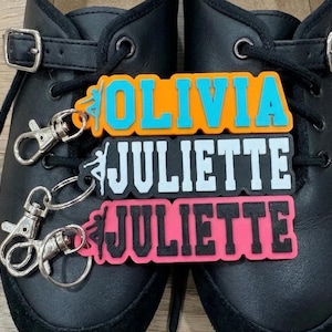 May include: Black shoes with buckles and laces, featuring three personalised keyrings. The top keyring is orange with "OLIVIA" in blue. The middle keyring is black with "JULIETTE" in white. The bottom keyring is pink with "JULIETTE" in black.