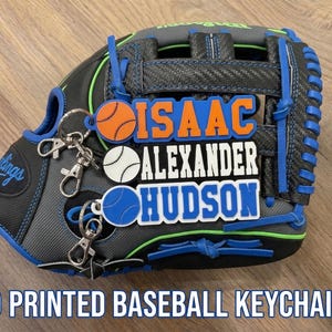 Könnte beinhalten: Drei 3D-gedruckte Baseball-Schlüsselanhänger mit den Namen ISAAC in Orange, ALEXANDER in Weiß und HUDSON in Blau. Jeder Schlüsselanhänger enthält ein kleines Baseball-Symbol und einen silbernen Karabinerhaken aus Metall, präsentiert auf einem schwarz-grauen Baseballhandschuh.