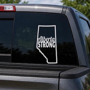 Puede incluir: Calcomanía de vinilo blanco con el contorno de Alberta, Canadá, y el texto "Alberta Strong" en una fuente apilada. La calcomanía está adherida a la ventana trasera de un vehículo oscuro, con una vista borrosa de árboles al fondo.