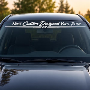 Puede incluir: Un adhesivo de vinilo blanco aplicado en la parte superior del parabrisas de un coche. El adhesivo muestra el texto "YOUR Custom Designed VINYL DECAL" en una fuente estilizada, subrayado por una línea horizontal con tres barras.