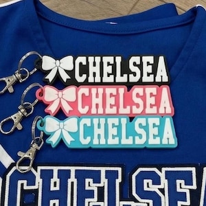 Può includere: Tre portachiavi rettangolari in silicone con un'icona a fiocco bianco sulla sinistra e il nome CHELSEA in lettere maiuscole bianche. I portachiavi sono neri, rosa e azzurri, ciascuno fissato a un moschettone in metallo argentato.