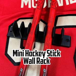May include: Black mini hockey stick wall rack holding two red hockey sticks. The sticks are positioned against a red jersey with white and black accents. The text "Mini Hockey Stick Wall Rack" is displayed in white.