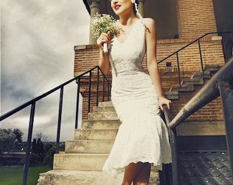 May include: A woman in a white lace dress and white heels stands on a set of stone steps. She is holding a bouquet of white flowers and looking up at the sky. The background is a brick building with arched doorways.