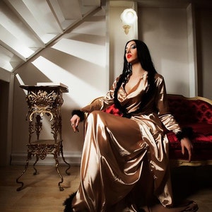 May include: A woman in a long, gold satin robe with black feather trim sits on a red velvet couch. She is wearing red lipstick and has long, dark hair. The couch is in a room with a gold decorative stand and white walls.