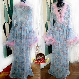 Puede incluir: Una bata larga de color azul claro con estampado floral y ribete de plumas rosas. La bata tiene mangas largas y un lazo en la cintura.
