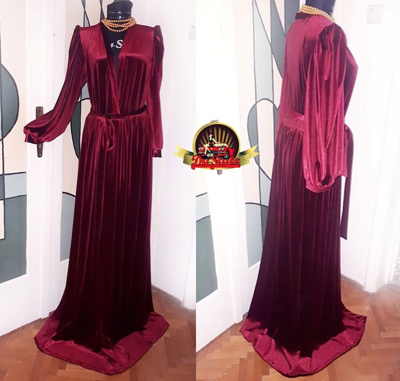May include: A long, burgundy velvet robe with a V-neckline, long sleeves, and a tie belt. The robe has a flowing, elegant silhouette.