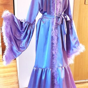 Peut inclure: Une longue robe de satin violette avec une bordure en plumes roses duveteuses. La robe a une ceinture à la taille et de longues manches avec des poignets bouffants.