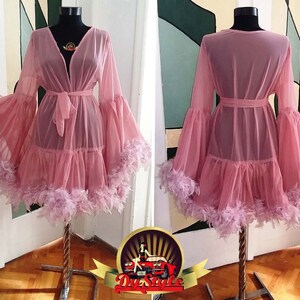 Puede incluir: Una bata de tul rosa con un dobladillo con volantes y ribete de plumas. La bata tiene mangas largas con pu&ntilde;os acampanados y un lazo en la cintura.