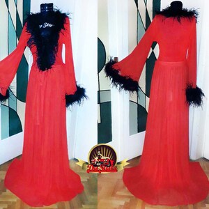 Puede incluir: Una bata roja de gasa transparente con ribete de plumas negras alrededor del cuello, las mangas y el dobladillo. La bata tiene una silueta larga y fluida y un lazo en la cintura.