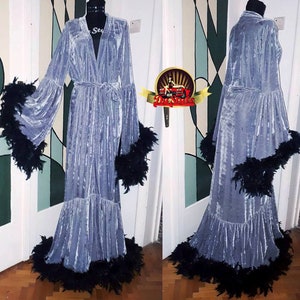 Puede incluir: Una larga bata de terciopelo azul claro con ribete de plumas negras alrededor de los puños, el dobladillo y el cuello. La bata tiene un cinturón de atar y un cuello en V.