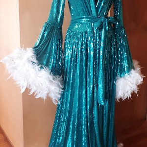 Könnte beinhalten: Eine türkisfarbene Paillettenrobe mit langen Ärmeln und einem weißen Federnbesatz. Die Robe hat einen Gürtel in der Taille und einen V-Ausschnitt.
