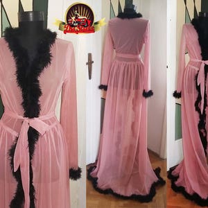 Op de afbeelding: Een doorschijnende roze badjas met zwarte verenbies. De badjas heeft een lange, vloeiende silhouet en een strik in de taille.