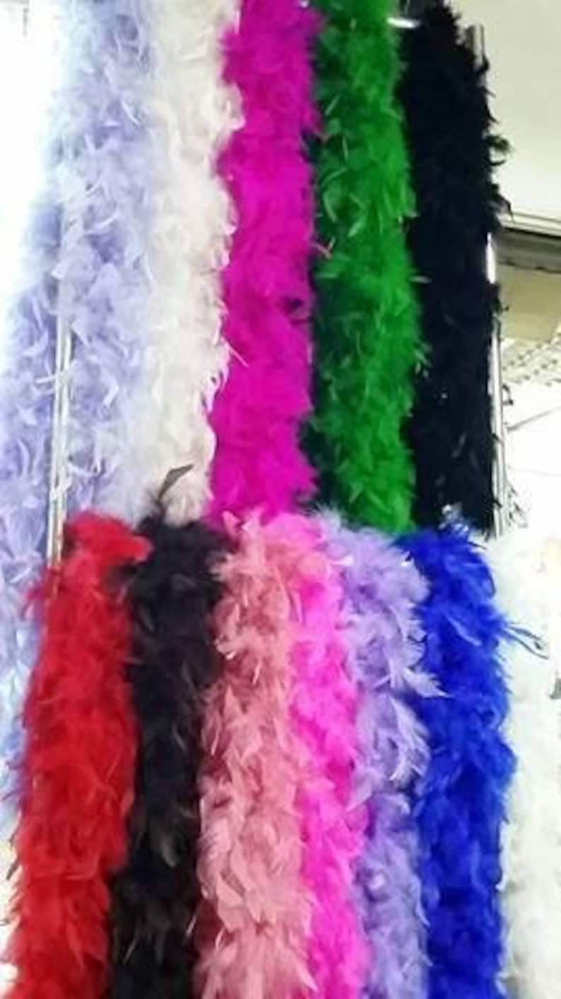 Puede incluir: Surtido de boas de plumas de colores en varios colores, incluyendo blanco, rosa, morado, rojo, negro y verde. Las boas est&aacute;n dispuestas verticalmente en un estante.