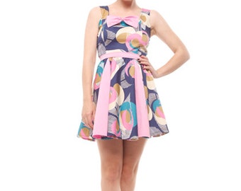 Peut inclure: Une femme portant une robe à motifs bleus et roses avec un nœud rose. Elle est debout, les jambes croisées, et porte des chaussures blanches.