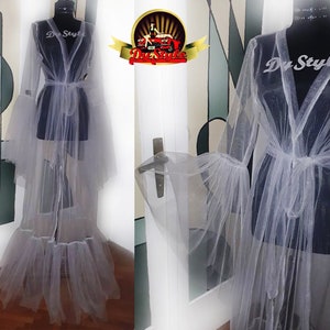 Puede incluir: Una bata de tul gris claro y transparente con mangas acampanadas y cinturón anudado. La bata se exhibe en un maniquí negro. El texto "Du Style" es visible en el maniquí. La bata tiene un dobladillo con volantes.