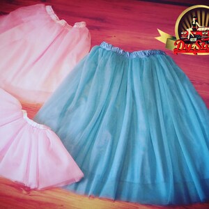 May include: Two tulle skirts, one light pink and one teal blue, are laid out on a wooden floor. The skirts are made of soft, flowing fabric and have a delicate, feminine look.