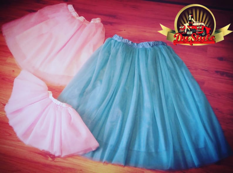 May include: Two tulle skirts, one light pink and one teal blue, are laid out on a wooden floor. The skirts are made of soft, flowing fabric and have a delicate, feminine look.