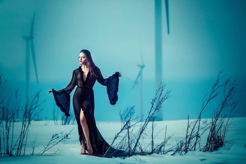 May include: A woman in a black sheer robe with a fur trim walks through a snowy field with wind turbines in the background.