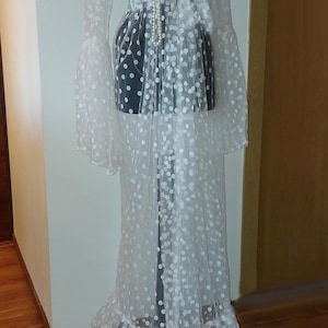 Puede incluir: Un vestido maxi blanco de tul transparente con lunares blancos y un dobladillo con volantes. El vestido tiene manga larga y un lazo en la cintura.