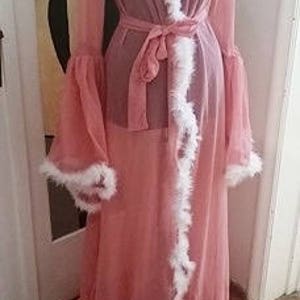 Puede incluir: Una larga bata rosa, transparente, con ribete de plumas blancas. La bata tiene una faja y mangas largas con puños con volantes.