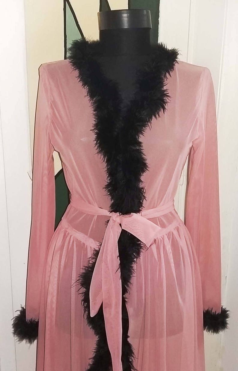 Op de afbeelding: Een doorschijnende roze badjas met zwarte verenbies rond de halslijn, mouwen en zoom. De badjas heeft een strik in de taille.