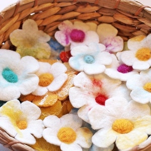 Op de afbeelding: Een rieten mand gevuld met viltbloemenversieringen. De bloemen zijn wit met verschillende gekleurde centra, waaronder geel, blauw en roze.