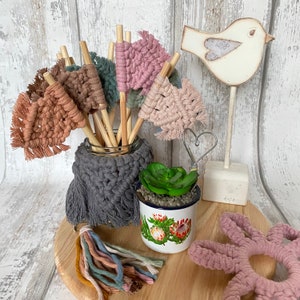 May include: A collection of colourful macrame wall hangings in pink, brown and grey, displayed in a glass jar with wooden sticks. The wall hangings are decorated with tassels and fringe. A small potted succulent plant and a wooden bird figurine are also included in the image.
