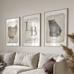 May include: A living room interior with a white sofa, a round wooden coffee table, a gray throw blanket, and three framed prints on the wall. The prints feature a map of Ohio, a map of Georgia, and a monogram with the initials "LB".
