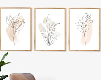 以下が含まれることがあります： 白い背景に植物のミニマルな線画が描かれた額装されたプリント3枚。それぞれのプリントには、ベージュとピーチの色合いの微妙な水彩のぼかしが施された、異なる植物が描かれています。