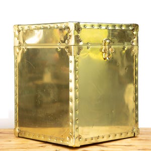 May include: A square brass storage box with a polished gold-tone finish. The box features reinforced metal corners, a series of decorative rivets along the edges, and a central metal latch mechanism on the front panel for secure closure.