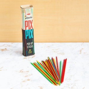 Peut inclure: Une boîte vintage en métal de bâtonnets de pick-up en plastique, appelée "Pix Pix", contenant 31 bâtonnets. La boîte est noire avec des rayures bleues, rouges et jaunes et les mots "Plastic Pix Pix Pick-Up Sticks 31 Plastic Sticks" imprimés dessus. Les bâtonnets sont éparpillés sur une surface en marbre blanc.
