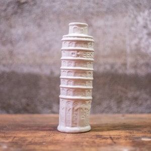 May include: White ceramic vase shaped like the Leaning Tower of Pisa. The vase has a textured surface and is approximately 25 cm tall.