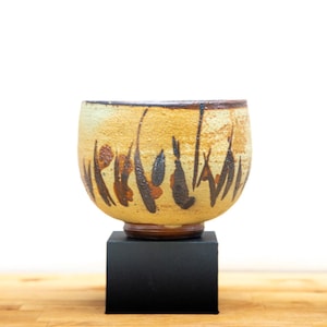 May include: A handcrafted ceramic bowl in beige and brown tones. The bowl has a distinctive pattern of dark brown and reddish-brown brushstrokes. It is presented on a small, black square plinth, highlighting its artistic design.