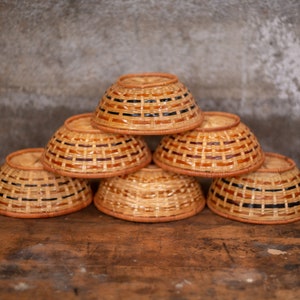May include: A stack of six woven brown and black bowls with a natural wood grain pattern. The bowls are stacked in a pyramid shape on a wooden surface.