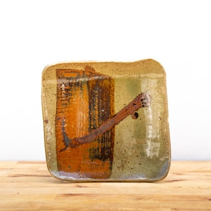 May include: Square ceramic dish with a unique, abstract design. The dish features a blend of colors, including orange, brown, and green, with a textured surface. A brown, curved accent adds visual interest. The dish is placed on a wooden surface.