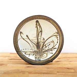 Puede incluir: Un plato redondo de cerámica con interior color crema y borde marrón oscuro. El plato presenta un diseño floral abstracto marrón. El plato se exhibe sobre una superficie de madera.