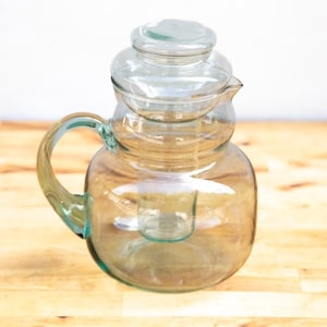 May include: A clear glass jug with a handle and a lid. The jug has a rounded body and a spout. The handle is curved and attached to the side of the jug. The lid sits on top of the jug. The jug is on a wooden surface.