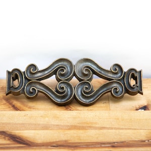 May include: Bronze-colored decorative hardware piece with a symmetrical design of swirling scrollwork. The hardware is set against a light wood surface and a white background. This piece could be used for furniture or other decorative applications.