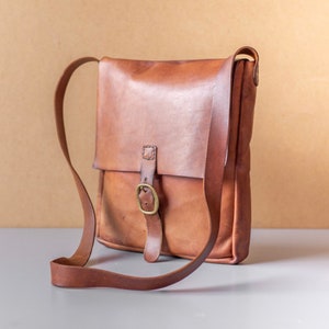 May include: A brown leather satchel bag with a buckle closure and a long adjustable strap. The bag is on a light gray surface.