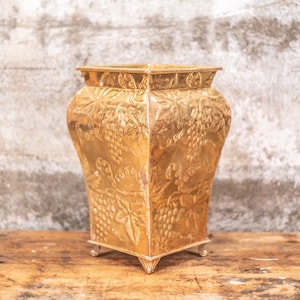 May include: A gold metal planter with a grapevine design. The planter has a square base and a rounded top. It is sitting on a wooden surface.
