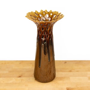 May include: A tall, brown ceramic vase with a glossy finish and a wide, flared, open-work rim. The upper section features a series of irregular, elongated cut-outs, while the lower body has a smooth, tapered shape with dark brown glaze drips.