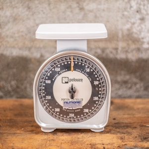Puede incluir: Una báscula de cocina vintage blanca con una cara negra y detalles plateados. La báscula tiene un dial circular con marcas para gramos y onzas. La báscula está etiquetada como "Pelouze Portion Controller Almore".