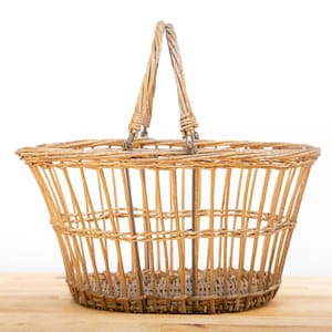 May include: A light brown wicker basket with a handle. The basket is oval-shaped with a woven design and a sturdy handle. The basket is sitting on a light brown wooden surface.