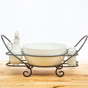 May include: A black metal serving caddy with scroll-style feet and handles, holding a large white ceramic bowl in the center, flanked by a white ceramic oil cruet on the left and two small white ceramic shakers on the right side of the frame.
