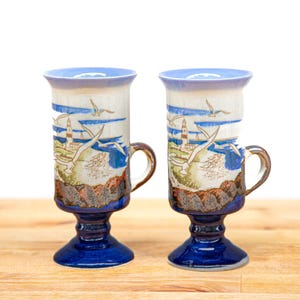 Op de afbeelding: Twee keramische latte mokken met een kustontwerp. De mokken hebben een blauwe voet, een bruin handvat en een uitlopende rand. Het ontwerp omvat een vuurtoren, zeemeeuwen en oceaangolven in tinten blauw, wit en bruin.