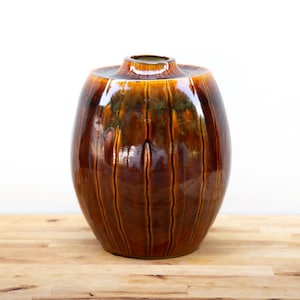 May include: A brown ceramic vase with vertical grooves and a glossy finish. The vase has a wide, rounded body and a narrow neck. The color transitions from dark brown to a lighter, amber hue. The vase is a decorative item.