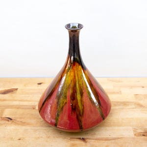 May include: A decorative ceramic vase with a teardrop shape. The vase features a glossy finish with vertical stripes of red, yellow, and brown. The vase has a narrow neck and a wide base, and it sits on a wooden surface.